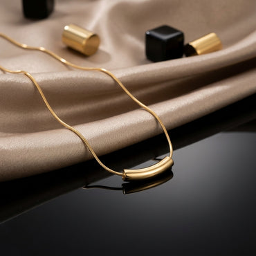 22K Gold-Tone Curved Tube Bar Pendant Snake Chain Necklace on black reflective surface with champagne satin fabric and black & gold geometric decorative pieces backdrop