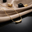 22K Gold-Tone Curved Tube Bar Pendant Snake Chain Necklace on black reflective surface with champagne satin fabric and black & gold geometric decorative pieces backdrop