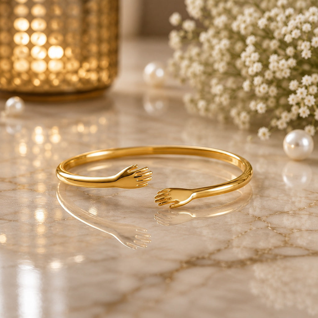 22K Gold-Tone Hug Me Hands Open Cuff Bangle Bracelet on cream marble surface with gold geometric candle holder, scattered white pearls and baby's breath flowers backdrop