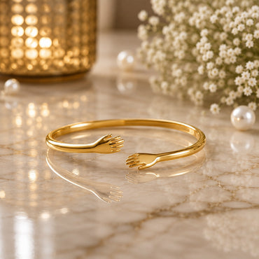 22K Gold-Tone Hug Me Hands Open Cuff Bangle Bracelet on cream marble surface with gold geometric candle holder, scattered white pearls and baby's breath flowers backdrop