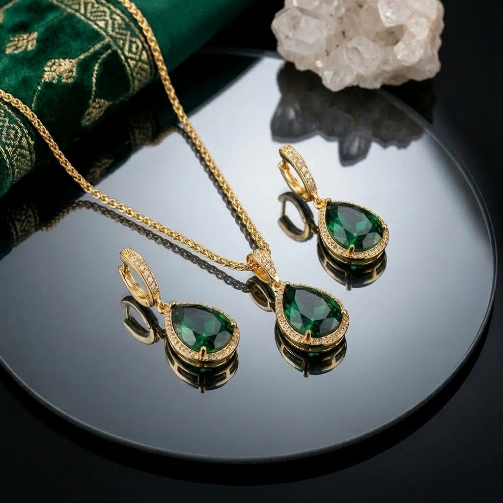 22K Gold-Tone Pear Emerald CZ Halo Pendant Necklace & Huggie Drop Earrings Jewelry Set flat lay on black mirror surface with emerald green embroidered fabric and white quartz crystal cluster backdrop