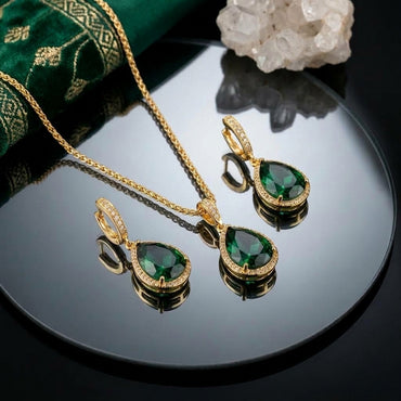 22K Gold-Tone Pear Emerald CZ Halo Pendant Necklace & Huggie Drop Earrings Jewelry Set flat lay on black mirror surface with emerald green embroidered fabric and white quartz crystal cluster backdrop