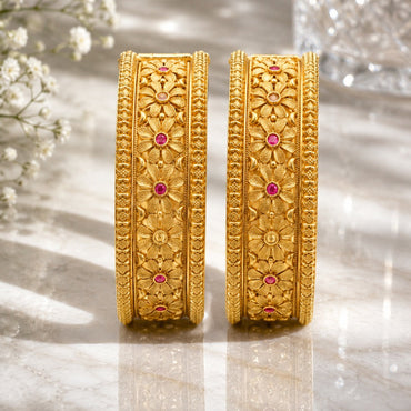 22K Gold-Tone Wide Embossed Floral Repousse & Ruby CZ Bridal Kada Set of 2 standing upright on marble surface with white baby's breath flowers and crystal glass vase backdrop