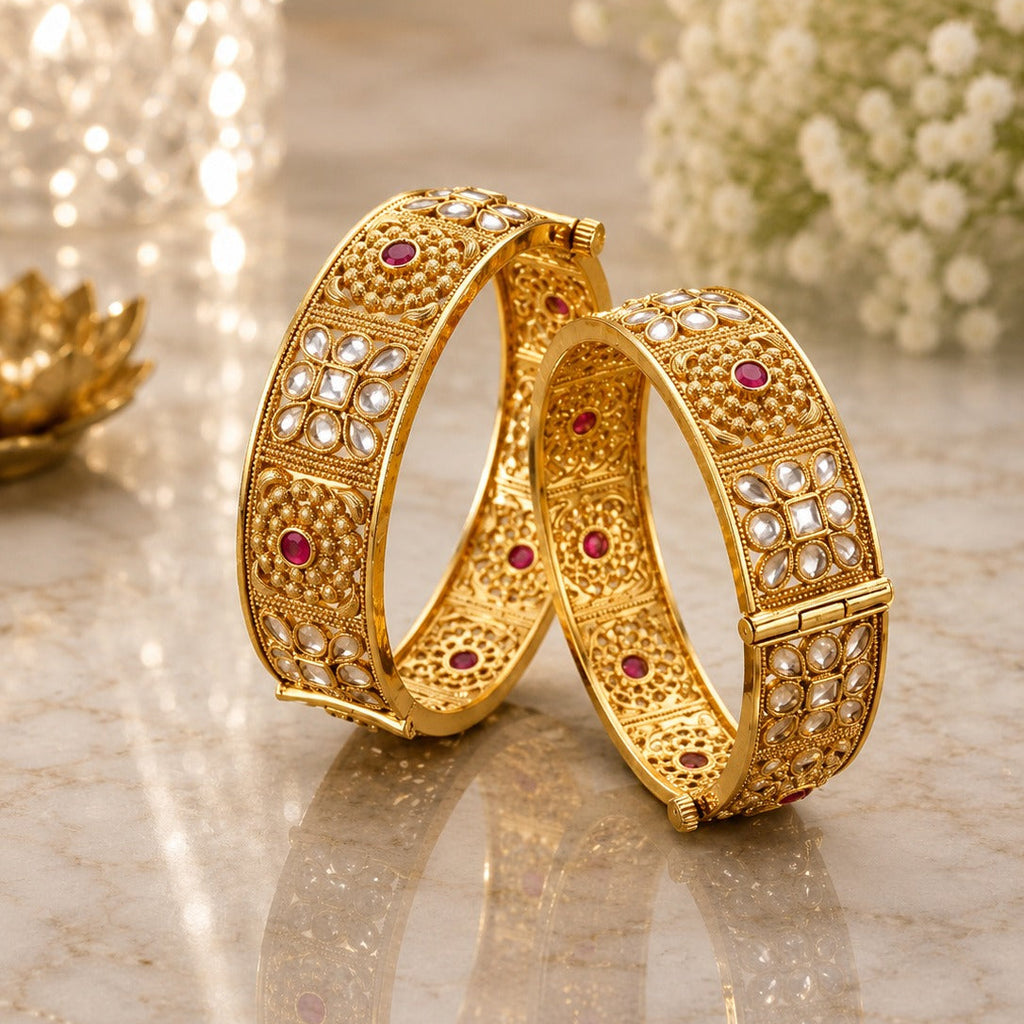 22K Gold-Tone Wide Filigree Kundan Flower & Ruby CZ Granule Bridal Kada Set of 2 on marble surface with gold lotus candle holder and white baby's breath flowers backdrop
