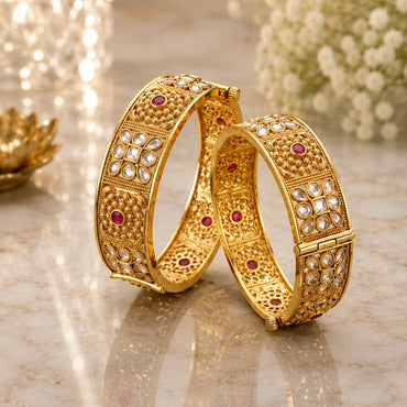 22K Gold-Tone Wide Filigree Kundan Flower & Ruby CZ Granule Bridal Kada Set of 2 on marble surface with gold lotus candle holder and white baby's breath flowers backdrop