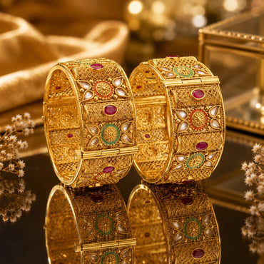 22K Gold-Tone Wide Filigree Kundan Ruby CZ & Meenakari Enamel Bridal Kada Set of 2 on dark reflective surface with gold jewelry box and dried flower sprigs backdrop