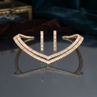 Gold-Tone Double-Row Baguette CZ V-Shape Collar Necklace and Bar Drop Earrings Set flat lay on dark grey mirror surface with teal velvet and burgundy silk backdrop showing rigid V-shape double-row baguette CZ collar necklace beside two slim vertical baguette CZ bar drop earrings with mirror reflections below