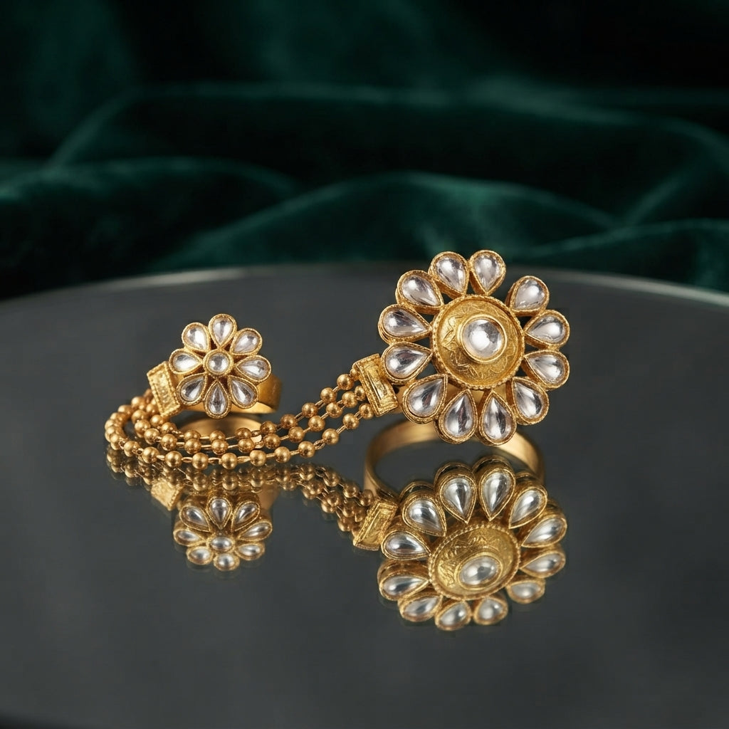Gold Kundan Polki Double Finger Chain Ring Sunflower Hath Phool — 22K antique gold-tone Mughal-inspired Kundan hath phool double finger ring with large sunflower daisy Kundan polki main ring of 8 to 10 teardrop petal stones connected via 3-strand gold ball chain to matching small 5-petal Kundan flower pinky ring, on dark grey mirror surface with emerald green crushed velvet luxury editorial backdrop
