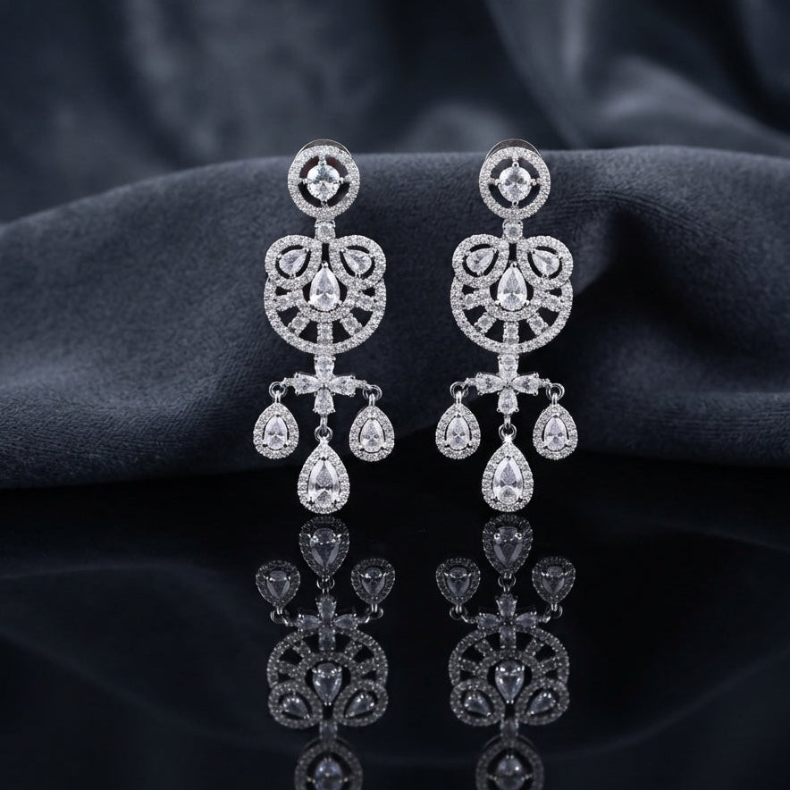 Silver CZ Medallion and Triple Pear Halo Chandelier Drop Earrings — rhodium silver-tone bridal chandelier earrings with round CZ medallion stud open Gothic arch chandelier body with trefoil pear CZ halos cruciform CZ connector and 3-drop triple pear CZ halo fringe displayed on dark black mirror surface with charcoal satin backdrop