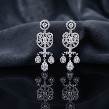 Silver CZ Medallion and Triple Pear Halo Chandelier Drop Earrings — rhodium silver-tone bridal chandelier earrings with round CZ medallion stud open Gothic arch chandelier body with trefoil pear CZ halos cruciform CZ connector and 3-drop triple pear CZ halo fringe displayed on dark black mirror surface with charcoal satin backdrop