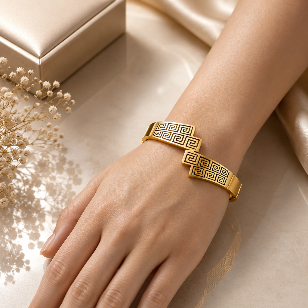 Gold-tone Greek key meander dual bar open cuff bangle worn on wrist showing intricate geometric engraving with dark antique fill on champagne satin background with dried white flowers
