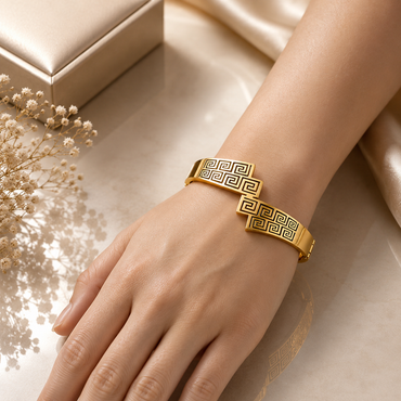 Gold-tone Greek key meander dual bar open cuff bangle worn on wrist showing intricate geometric engraving with dark antique fill on champagne satin background with dried white flowers