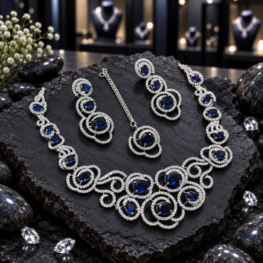 Royal blue oval and clear-accent jewellery set with a silver-tone swirl necklace, two drop earrings and matching chain maang tikka on dark slate.