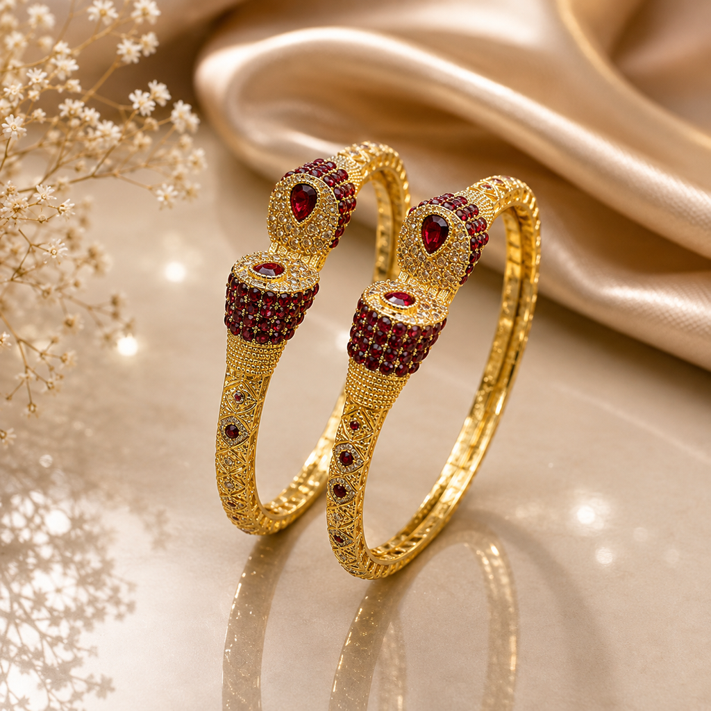 Set of two ruby red CZ and gold-tone floral dome engraved bangle bracelets with pear-cut ruby red teardrop crown stone geometric lattice band and scattered CZ accents displayed on champagne satin with dried white flowers