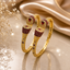 Set of two ruby red CZ and gold-tone floral dome engraved bangle bracelets with pear-cut ruby red teardrop crown stone geometric lattice band and scattered CZ accents displayed on champagne satin with dried white flowers