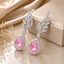 Silver-tone pink CZ teardrop and marquise leaf wing drop earrings featuring large pear-cut blush pink CZ center stone in round CZ halo frame with sweeping marquise and round CZ pave leaf wing top displayed on champagne satin with dried white flowers