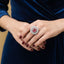 Person wearing a blue dress with a ring featuring a red gemstone, set against a blurred background.
