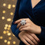 Person wearing a blue dress with a close-up of a hand holding a decorative ring against a blurred light background.