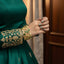 Green traditional outfit with gold embroidery and jewelry against a neutral background