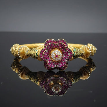 Gold bracelet with a floral design featuring red and green gemstones on a reflective surface.