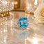 Silver-Tone Aqua Blue Emerald-Cut CZ Double Halo Cocktail Statement Ring product shot on marble surface with white rose and floral backdrop