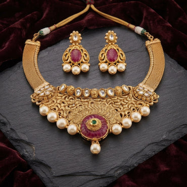 Antique Gold Ruby Pearl Mughal Filigree Bridal Choker Necklace Earrings Set on dark slate stone surface with burgundy velvet fabric backdrop showing wide mesh jali antique gold bib choker with dense filigree arabesque scroll work top row of oval polki uncut CZ stones large ruby garnet cabochon centre pendant with emerald centre faux pearl drop and cascading round faux pearl drops along base with matching gold filigree chandbali earrings featuring oval ruby CZ cabochon and four faux pearl drops