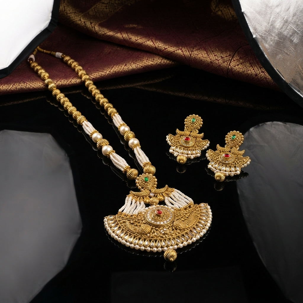 Antique Gold Ruby Pearl Mughal Rani Haar Long Necklace Earrings Set on black mirror surface with burgundy gold brocade backdrop showing double-strand gold ball bead and seed pearl chain with fan filigree pendant ruby red CZ emerald green CZ seed pearl fringe gold tassel drops and matching temple jhumka earrings