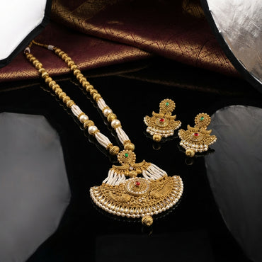 Antique Gold Ruby Pearl Mughal Rani Haar Long Necklace Earrings Set on black mirror surface with burgundy gold brocade backdrop showing double-strand gold ball bead and seed pearl chain with fan filigree pendant ruby red CZ emerald green CZ seed pearl fringe gold tassel drops and matching temple jhumka earrings