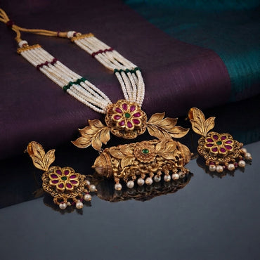 Antique Gold Temple Dabba Pendant Seed Pearl Necklace Earrings Set on dark grey mirror surface with purple and teal silk fabric backdrop showing 5-strand white seed pearl chain with silk knot spacers and large antique gold temple cylinder dabba pendant with ruby red kundan CZ floral top emerald green CZ scrollwork peacock motifs and faux pearl drop fringe alongside matching antique gold floral disc stud earrings with ruby red pear kundan CZ petals and pearl drops
