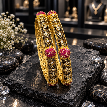 Two antique-style gold-tone South Indian kangans with floral scroll openwork, clear round accents, raised deep pink clusters and textured borders.