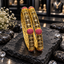 Two antique-style gold-tone South Indian kangans with floral scroll openwork, clear round accents, raised deep pink clusters and textured borders.