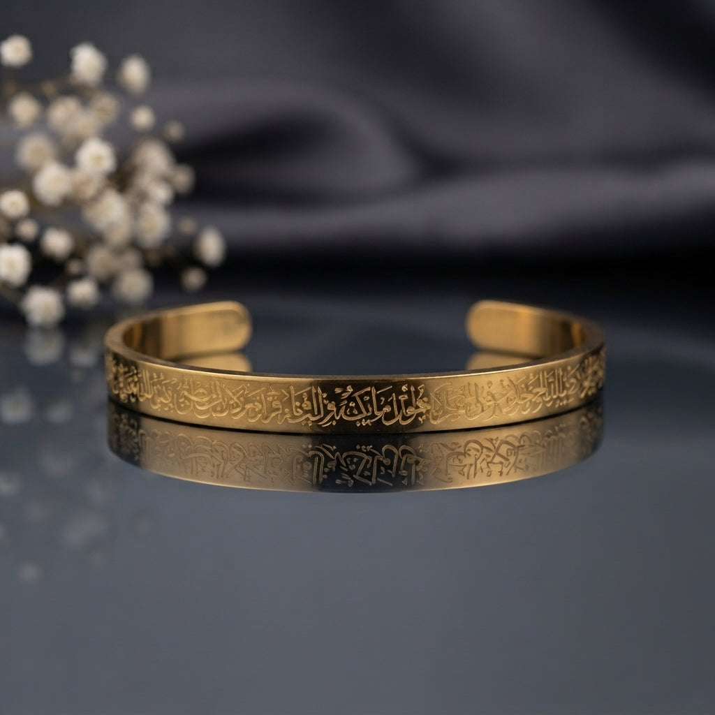 Ayat ul Kursi Arabic Calligraphy Cuff Bangle in 18K Gold-tone PVD stainless steel on dark mirror surface with white gypsophila flowers and grey silk backdrop showing slim open C-shape cuff bangle with continuous laser-etched Arabic Quranic calligraphy running around full circumference with mirror reflection below