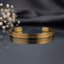 Ayat ul Kursi Arabic Calligraphy Cuff Bangle in 18K Gold-tone PVD stainless steel on dark mirror surface with white gypsophila flowers and grey silk backdrop showing slim open C-shape cuff bangle with continuous laser-etched Arabic Quranic calligraphy running around full circumference with mirror reflection below