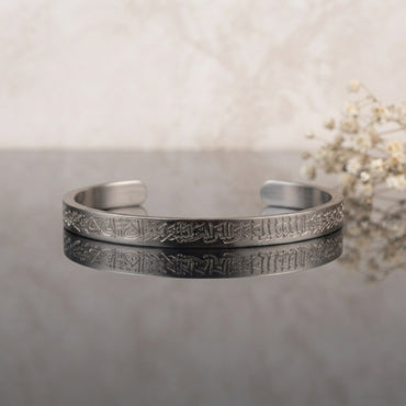 Ayat ul Kursi Arabic Calligraphy Cuff Bangle in Silver polished stainless steel on grey mirror surface with white gypsophila flowers backdrop showing slim open C-shape cuff bangle with continuous laser-etched Arabic Quranic calligraphy running around full circumference with mirror reflection below