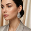Baguette CZ Teardrop Stud Earrings with Round Pavé Border in gold-tone plating worn on model ear with beige silk curtain backdrop and champagne gold blazer showing tiered baguette CZ teardrop with pavé round CZ border
