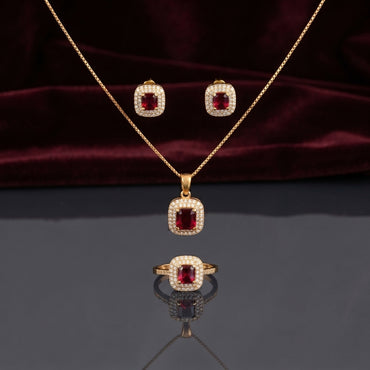 CZ Cushion Halo Pendant Necklace Stud Earrings and Ring Set in Ruby Red CZ gold-tone flat lay on burgundy wine velvet backdrop showing 3-piece set with deep ruby red cushion CZ center in white CZ micro-pavé halo gold-tone pendant necklace matching stud earrings and cocktail ring with mirror reflection