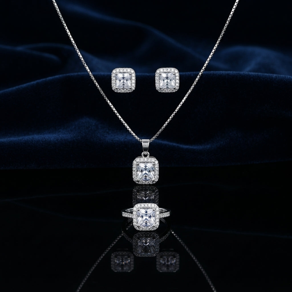 CZ Cushion Halo Pendant Necklace Stud Earrings and Ring Set in White CZ Diamond Illusion silver-tone flat lay on deep navy blue satin backdrop showing 3-piece set with all-white princess CZ center in white CZ micro-pavé halo pendant necklace matching stud earrings and cocktail ring with mirror reflection