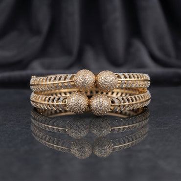 CZ Pave Ball Charm Multi-Row Open-Work Spine Bangle Pair in gold-tone finish displayed stacked on dark grey marble mirror surface with black silk drape backdrop showing two matching bangles with double-row chevron herringbone spine-rib lattice band continuous round CZ micro-pave and 4 large round CZ pave ball charms at center front with mirror reflection below