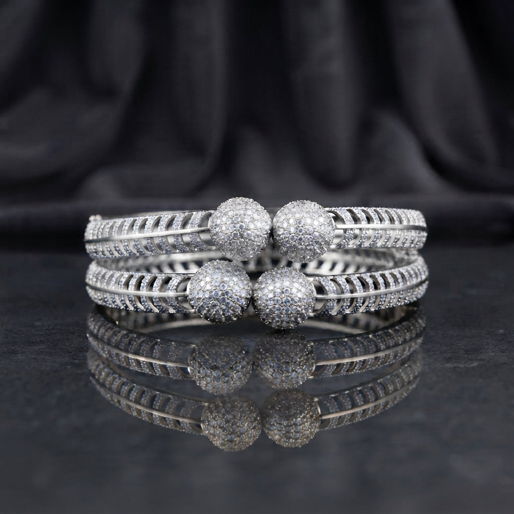CZ Pave Ball Charm Multi-Row Open-Work Spine Bangle Pair in silver rhodium-tone finish displayed stacked on dark grey marble mirror surface with black silk drape backdrop showing two matching bangles with double-row chevron herringbone spine-rib lattice band continuous round CZ micro-pave and 4 large round CZ pave ball charms at center front with mirror reflection below