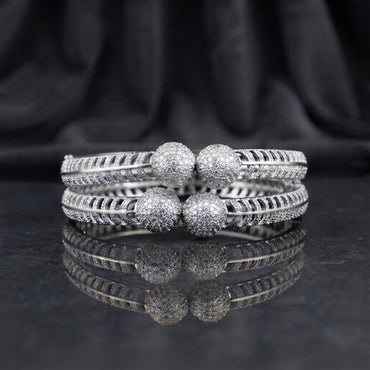 CZ Pave Ball Charm Multi-Row Open-Work Spine Bangle Pair in silver rhodium-tone finish displayed stacked on dark grey marble mirror surface with black silk drape backdrop showing two matching bangles with double-row chevron herringbone spine-rib lattice band continuous round CZ micro-pave and 4 large round CZ pave ball charms at center front with mirror reflection below