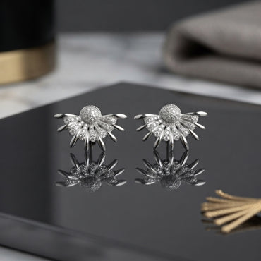 CZ Pave Sunburst Fan Stud Earrings in Silver pair on dark grey marble surface with gold accent props showing rhodium-plated bold statement studs with fully pave CZ domed ball centre and 10-12 slim tapered rays with CZ-filled fan body with mirror reflection below