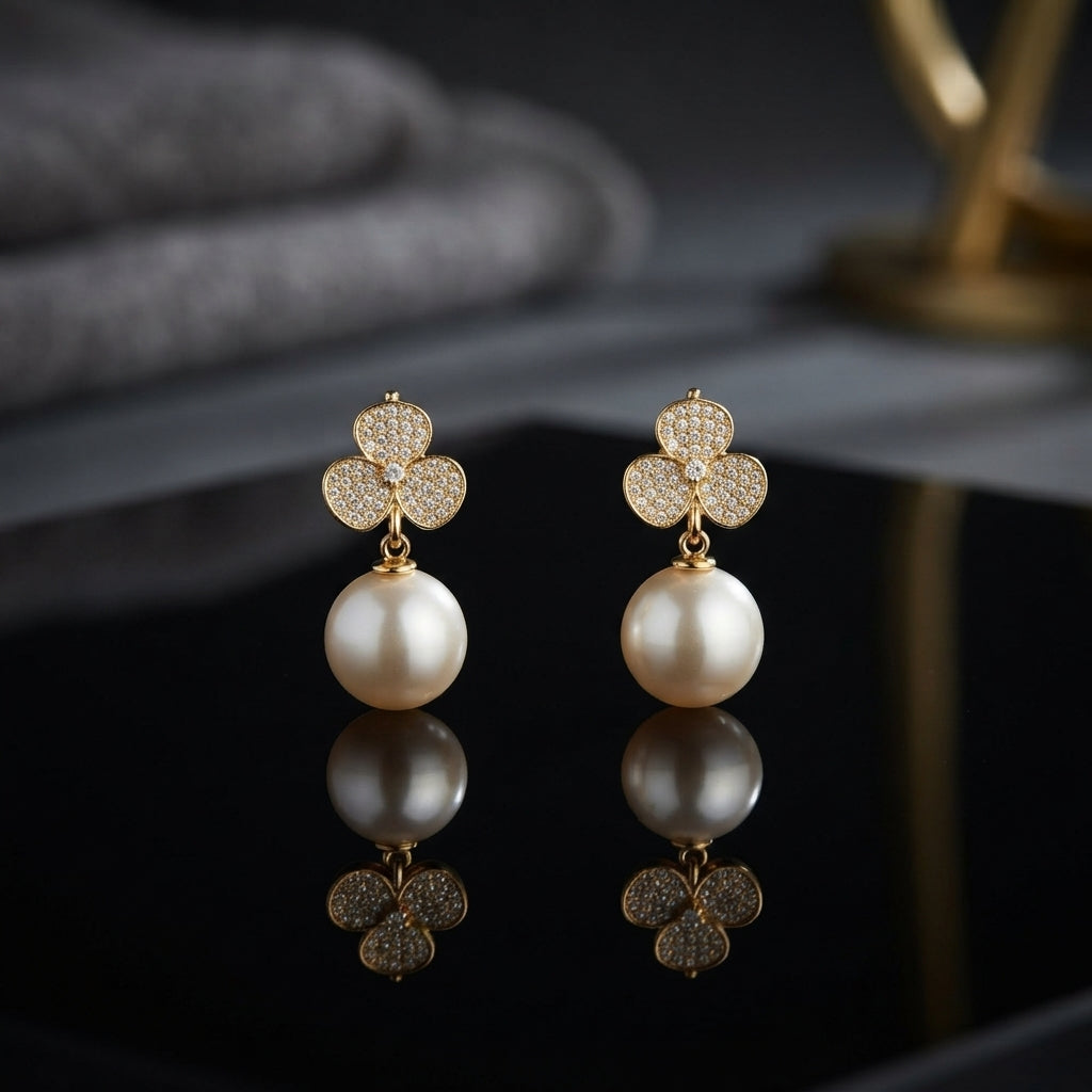 CZ Pave Trefoil Clover Pearl Drop Earrings in Gold pair on black surface with dark botanical backdrop and gold prop showing 18K gold-tone fully pave CZ 3-petal clover stud suspending large lustrous round white pearl drop with mirror reflection below
