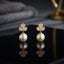 CZ Pave Trefoil Clover Pearl Drop Earrings in Gold pair on black surface with dark botanical backdrop and gold prop showing 18K gold-tone fully pave CZ 3-petal clover stud suspending large lustrous round white pearl drop with mirror reflection below