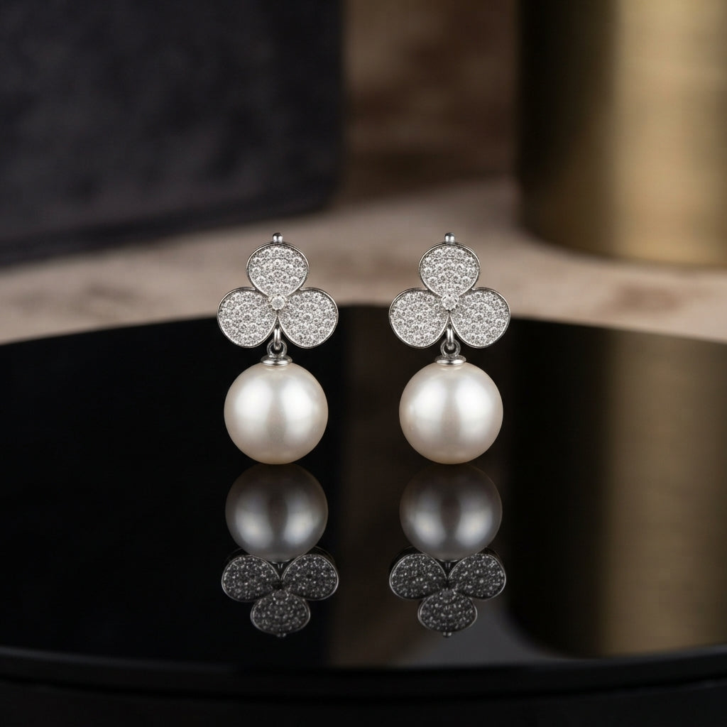 CZ Pave Trefoil Clover Pearl Drop Earrings in Silver pair on black round tray with warm gold prop backdrop showing rhodium-plated fully pave CZ 3-petal clover stud suspending large lustrous round white pearl drop with mirror reflection below