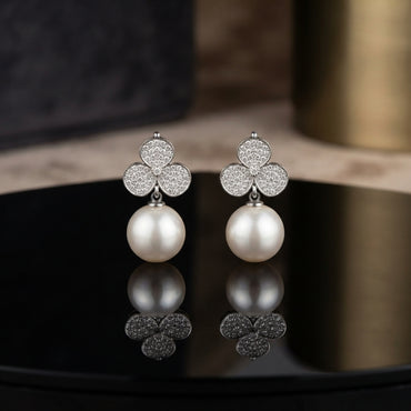 CZ Pave Trefoil Clover Pearl Drop Earrings in Silver pair on black round tray with warm gold prop backdrop showing rhodium-plated fully pave CZ 3-petal clover stud suspending large lustrous round white pearl drop with mirror reflection below
