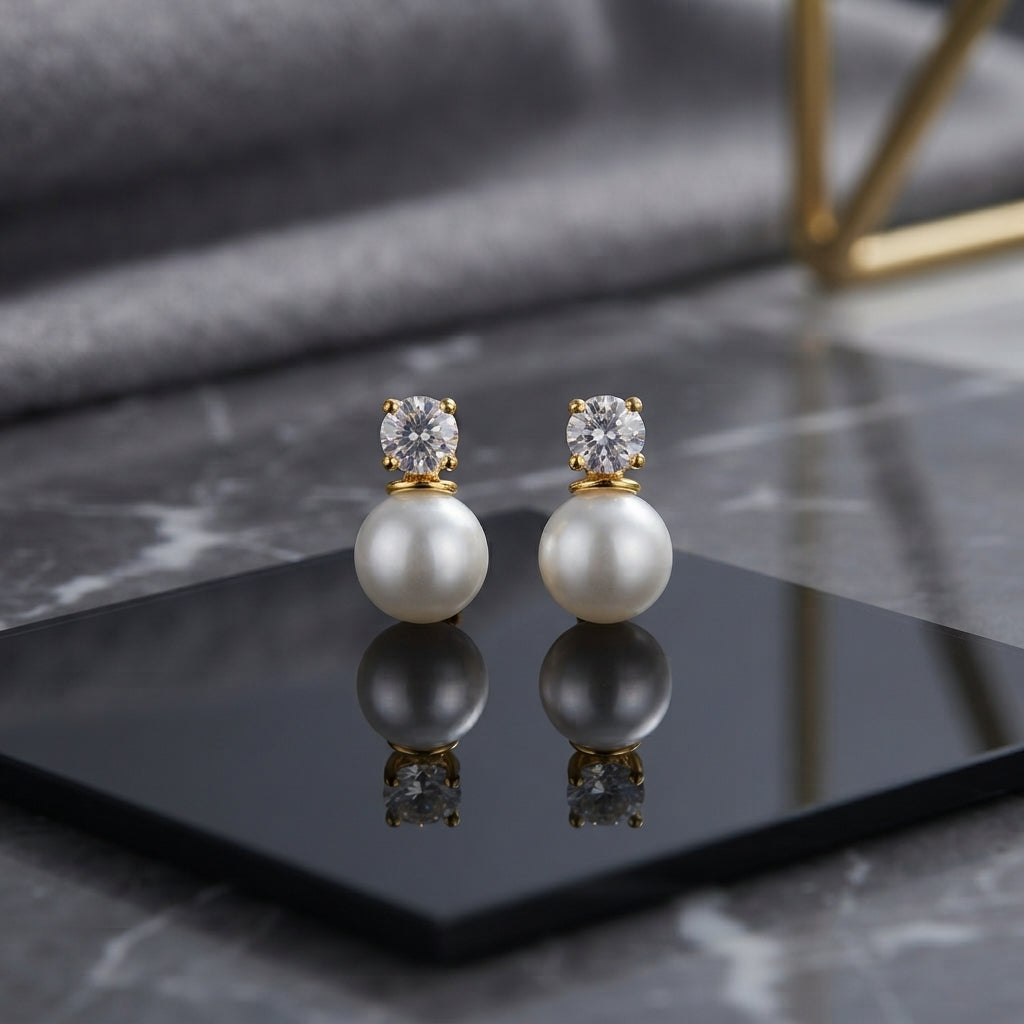 CZ Solitaire Pearl Drop Stud Earrings in Gold pair on black hexagonal tray with grey marble backdrop showing 18K gold-tone round brilliant CZ solitaire in 4-prong setting above large lustrous round white pearl with mirror reflection below