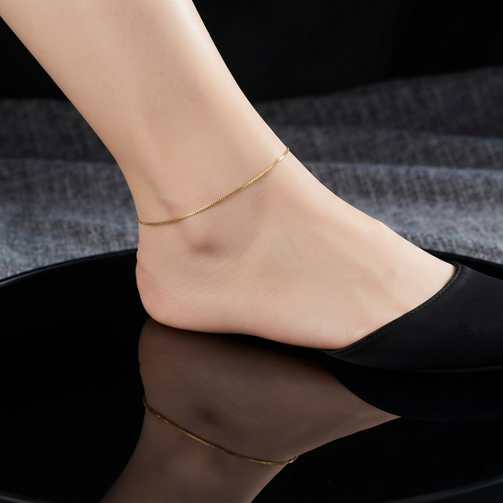 Dainty Gold Chain Anklet in 18K gold-tone worn on bare ankle with black pointed heel in editorial model shot showing ultra-slim minimalist fine box chain delicately wrapped around ankle with mirror reflection below on black lacquer surface