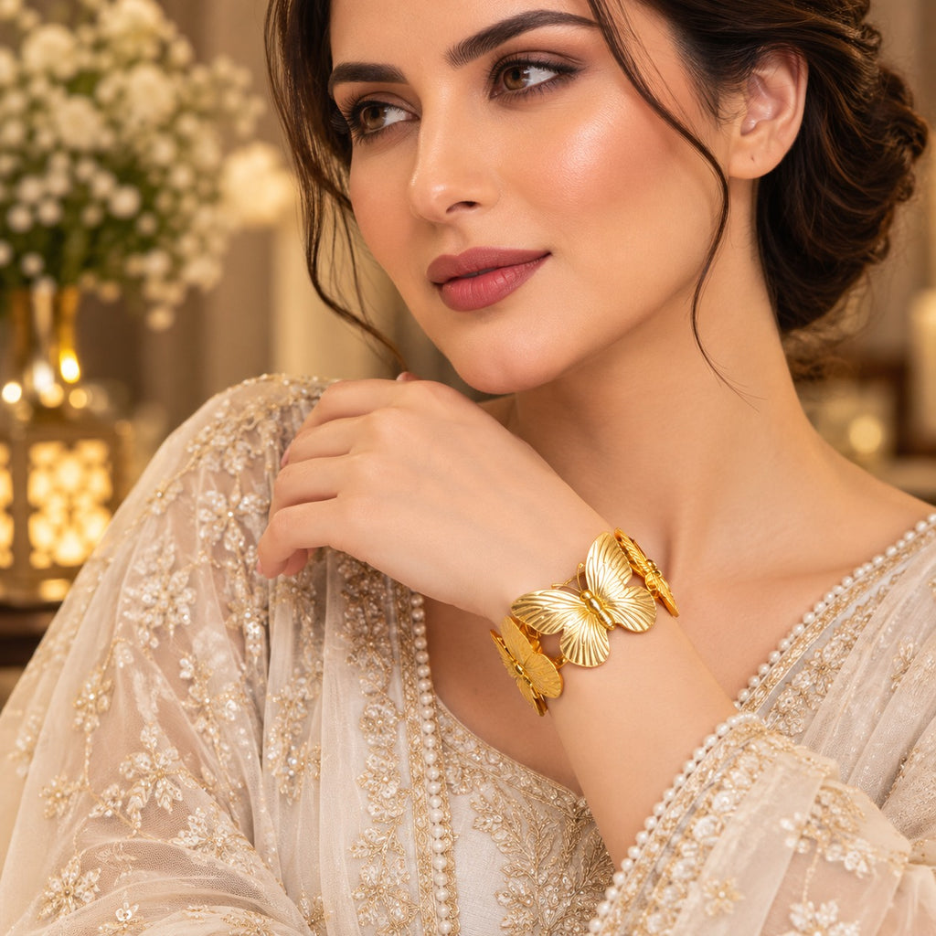 Gold 3D Butterfly Statement Cuff Bangle Bracelet — 18-22K gold-tone wide open cuff bangle with large sculpted 3D butterfly centerpiece featuring fine ribbed wing detailing in mirror-polish gold worn by model in ivory embroidered dupatta in luxury golden bokeh editorial setting