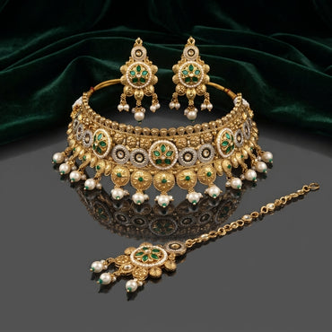 Gold Antique Emerald Green Kundan and Pearl Choker Bridal Set flat lay on dark grey mirror surface with emerald green velvet backdrop showing 3-piece set with wide kundan choker necklace featuring alternating emerald green 4-petal marquise kundan medallions on white enamel base with antique gold filigree fringe and triple pearl drops with matching chandelier earrings and maang tikka with mirror reflection