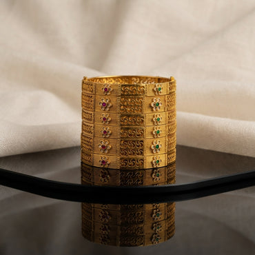Gold Antique Filigree Scroll Floral Ruby Emerald Bangles Set of 4 in 22K antique gold-tone plating on dark grey mirror surface with ivory cream fabric backdrop showing four wide rigid bangles stacked together with 3-panel band design featuring dense gold granulation panels with 4-petal flower motifs in ruby red and emerald green cabochon centres flanking open gold filigree swirl scroll arabesque centre panels with mirror reflection below